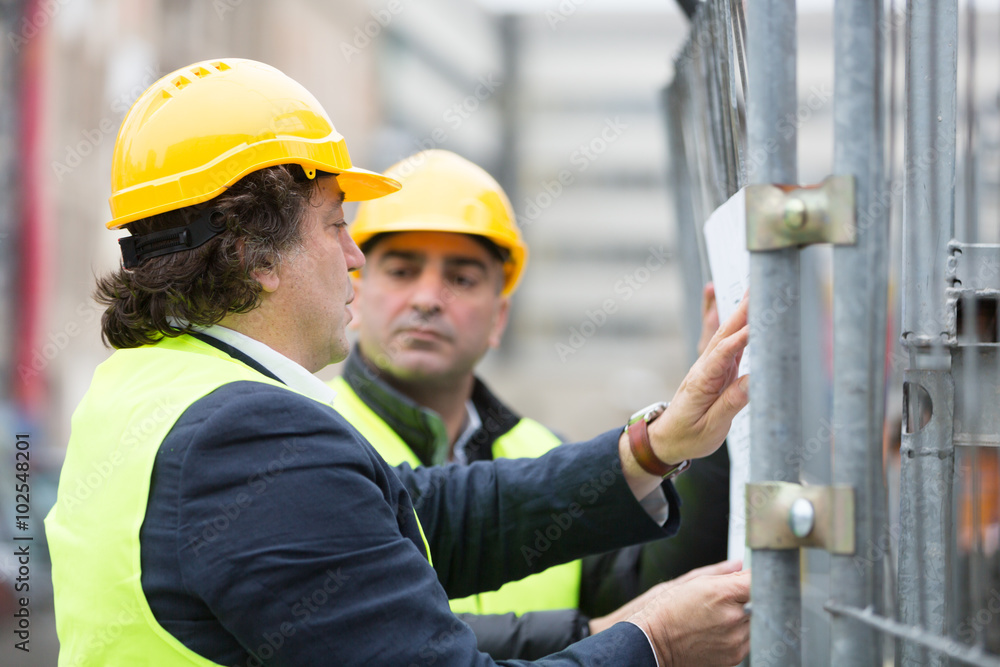 Two engineers wearing hardhat and yellow jacket reading office blueprints against scaffolding at construction site Two engineers wearing hardhat and yellow jacket reading office blueprints against scaffolding at construction site