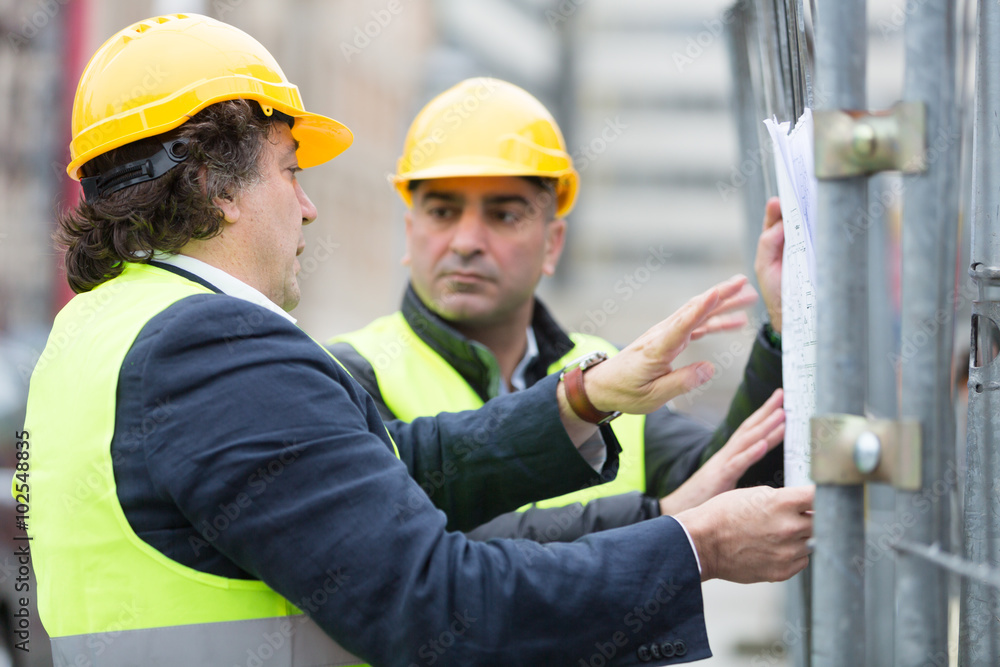Technicians with hardhat and yellow jacket reading office blueprints against scaffolding at construction site Technicians with hardhat and yellow jacket reading office blueprints against scaffolding at construction site