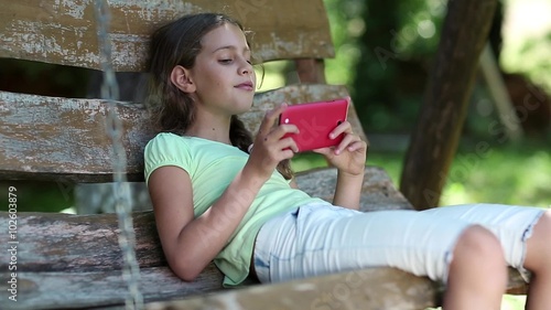 Girl with red smartphone sits on a swing bench in the garden and listens to music. Attractive girl uses smartphone. Beautiful girl with red smartphone sits on the swing bench photo