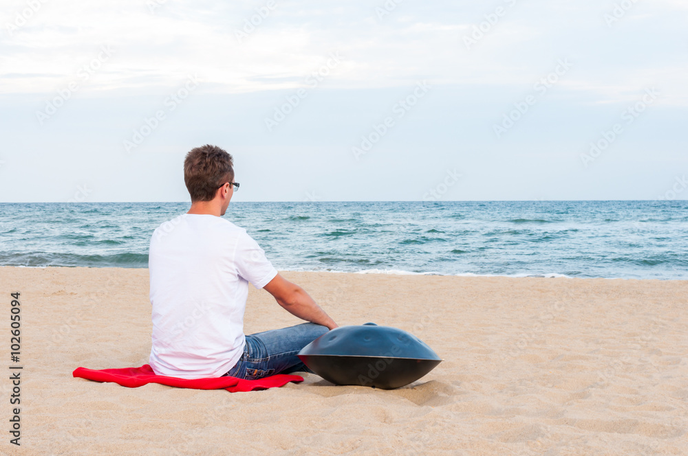 Young stylish guy sitting on the sand beach near handpan or hang with sea On Background. The Hang is traditional ethnic drum musical instrument