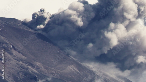 An aerial view of the Tungurahua volcano erupting in Ecuador, dense gray and white smoke billowing from the crater above the rugged barren slopes.