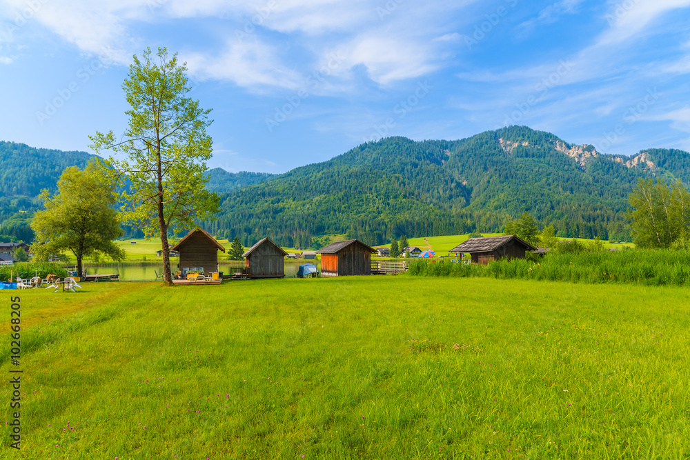 Green meadow in small alpine village on shore of Weissensee lake with traditional boat houses in background, Austria