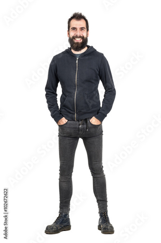 Laughing bearded punker in black hoodie with hands in pockets looking at camera. Full body length portrait isolated over white studio background. 