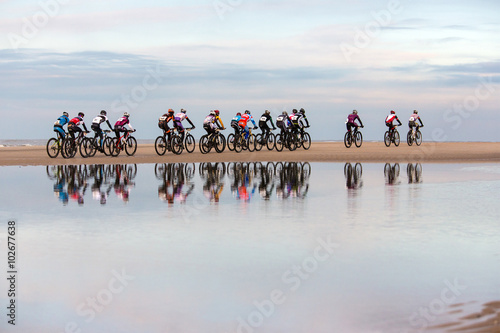 unrecognizable people drive with mountain bikes on a beach during a contest. with a visible reflection in the water in the foreground. 