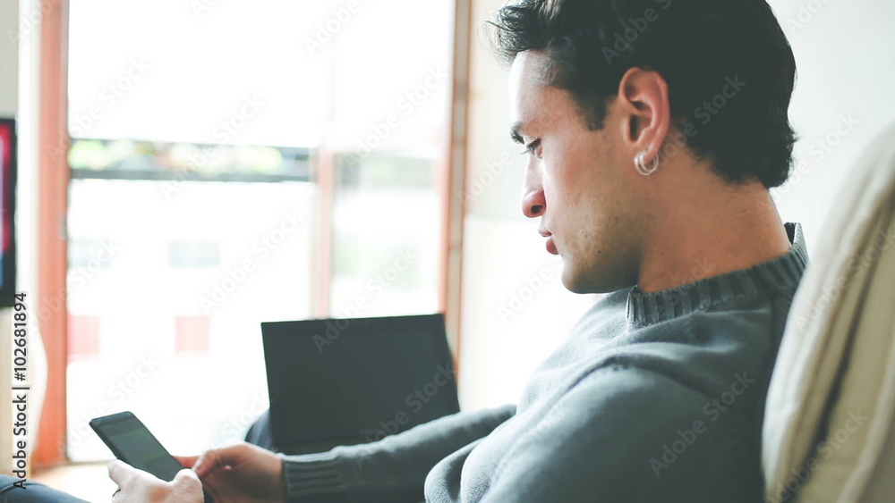 Rear view of young handsome caucasian brown hair man sitting on the sofa holding a smartphone tapping the screen with laptop on his knee - communication, happiness, social network concept Rear view of young handsome caucasian brown hair man sitting on the sofa holding a smartphone tapping the screen with laptop on his knee - communication, happiness, social network concept