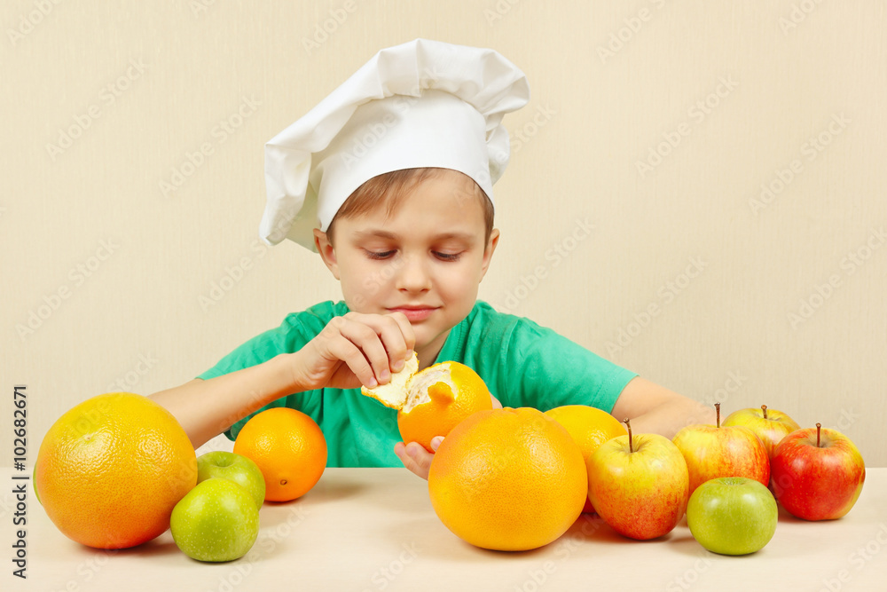 Little funny boy peeling fresh orange at the table with fruits Little funny boy peeling fresh orange at the table with fruits