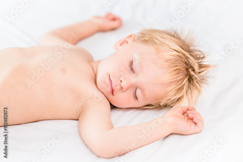 Close-up portrait of little boy with blond hair sleeping on white bed. Carefree childhood concept