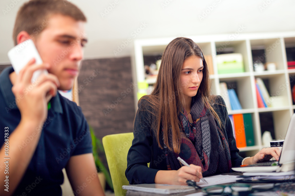 Beautiful smiling young businesswoman sitting working with a young man in a busy office. Man using a smart phone Beautiful smiling young businesswoman sitting working with a young man in a busy office. Man using a smart phone
