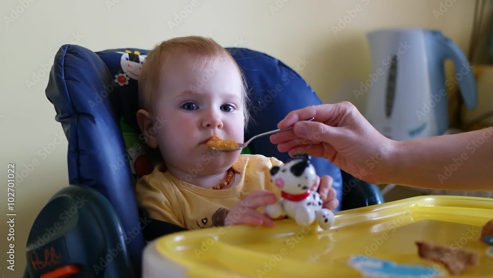 Baby sits in high char as mother feeds the child. Baby eats Baby sits in high char as mother feeds the child. Baby eats