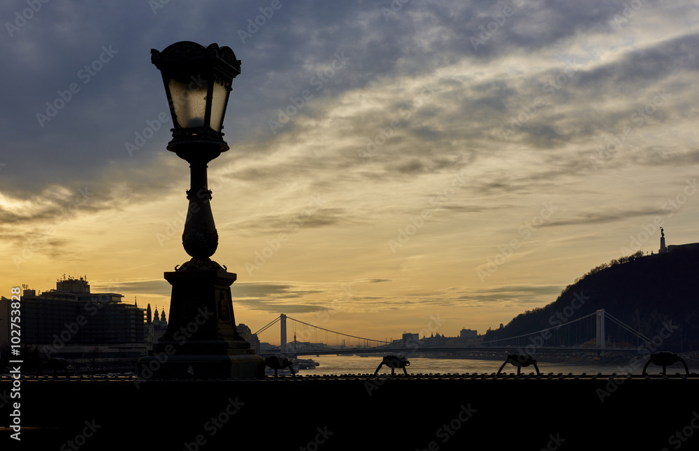 Lamp post in silhouette over Szechenyi Chain Bridge with dramatic sky in the background. Budapest, Hungary.