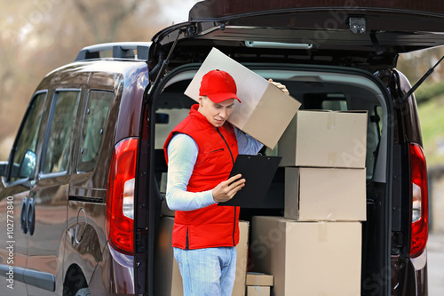 Young handsome delivery man standing near the car with boxes and packages, outdoors