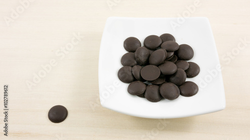 The group of dark chocolate buttons and small white square disk on light brown wooden board.