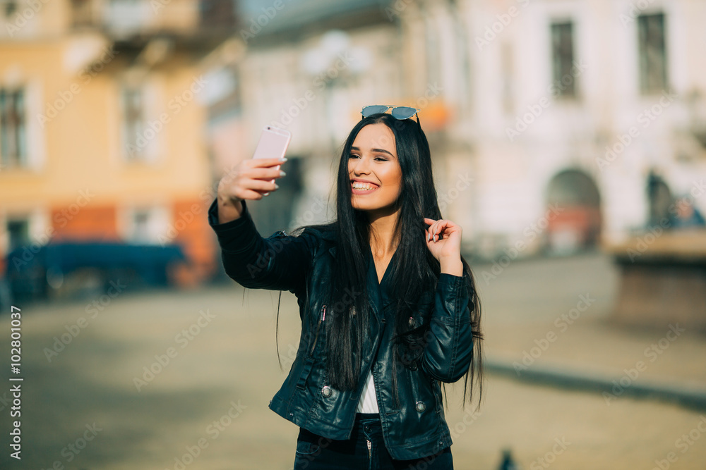 Fototapeta premium Young beautiful casual girl takes a selfie on her cell phone with sunglasses in jacket an black bag smile to camera on spring city street. Spring outfit, Spring street look.