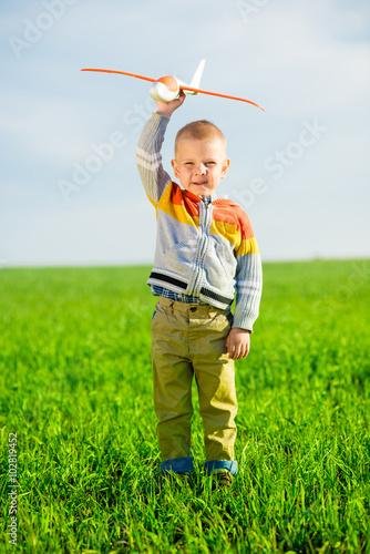Happy boy playing with toy airplane against blue summer sky and green field background.