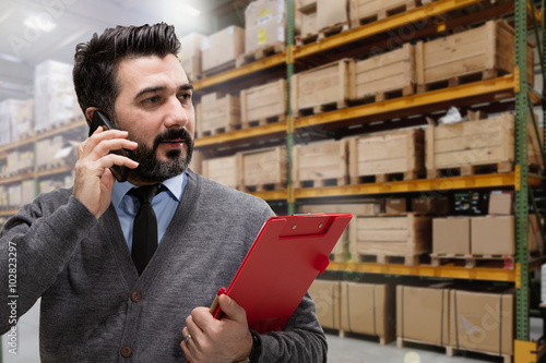 Corporate businessman using his cellphone while checking inventory in a large warehouse