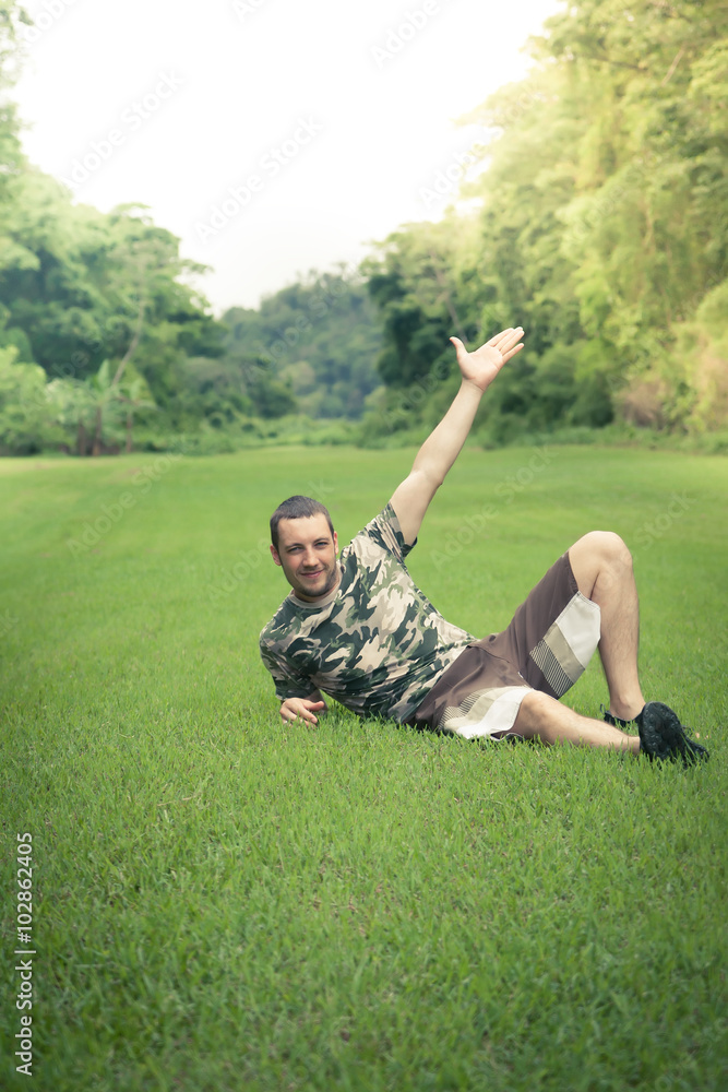 Young smiling man lying down on lawn in park with outstretched arm pointing to the sky