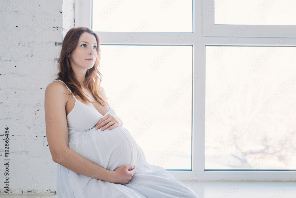 pregnant red-haired girl at the window in a white dress hugging belly pregnant red-haired girl at the window in a white dress hugging belly