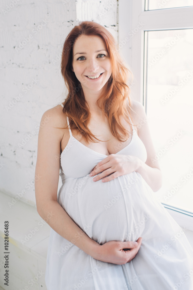 happy pregnant red-haired girl at the window in a white dress hugging belly happy pregnant red-haired girl at the window in a white dress hugging belly