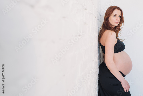 Pregnant woman with red hair in black clothes on a white wall © lumenphotos