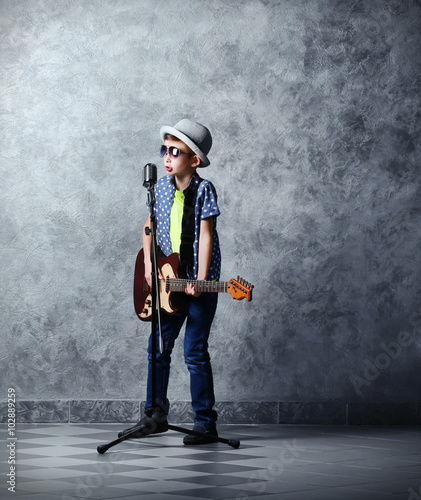 Little boy playing guitar and singing with microphone on a grey wall background © Africa Studio