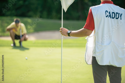 Golf caddy holding a flag for putting, golfer in the background reading green photo