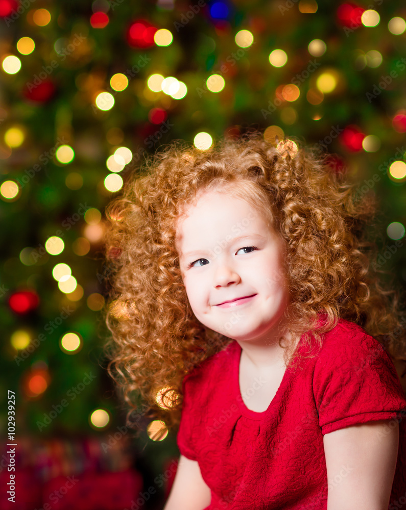 Pretty red-haired little girl wearing red dress sitting in front of Christmas tree with colorful lights Pretty red-haired little girl wearing red dress sitting in front of Christmas tree with colorful lights