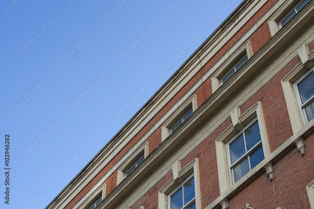 The facade of an old, red brick building with blue sky above. The colonnade of a period building with rows of uniform windows. A view from the ground looking upwards. The facade of an old, red brick building with blue sky above. The colonnade of a period building with rows of uniform windows. A view from the ground looking upwards.