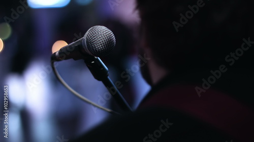 Close-up picture of singer sitting toward a microphone at the stage. Bearded man sitting back to the camera before the show starts.