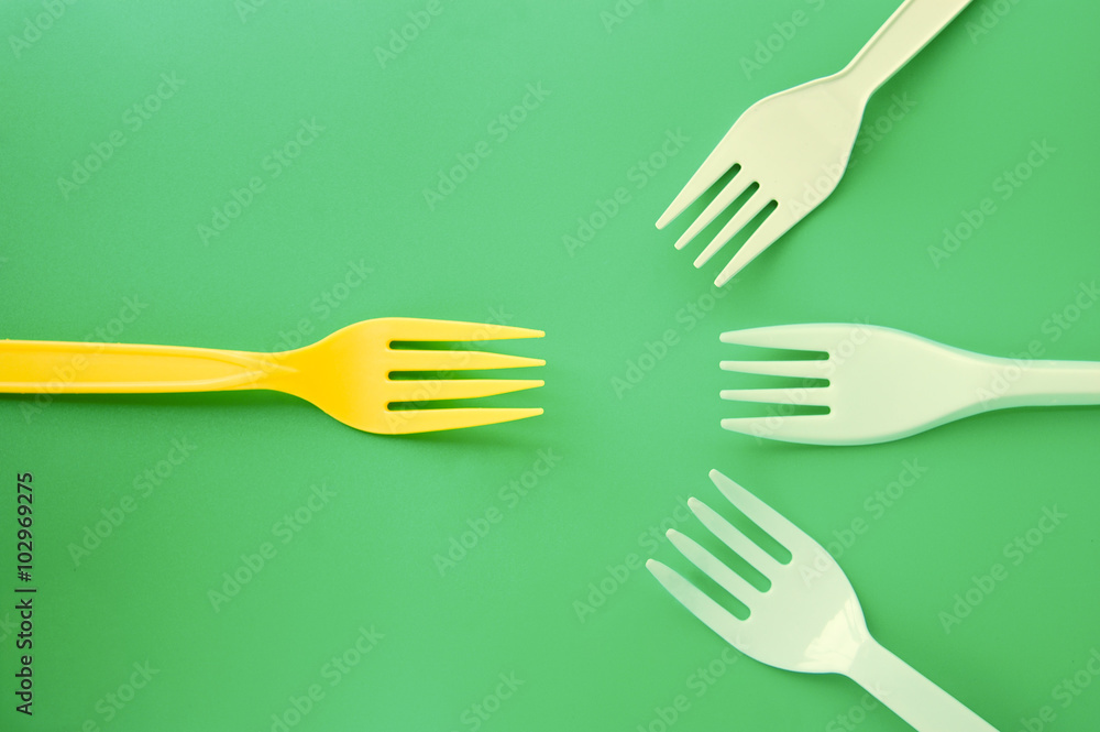 White plastic forks appearing to come together to battle yellow fork on a green background White plastic forks appearing to come together to battle yellow fork on a green background