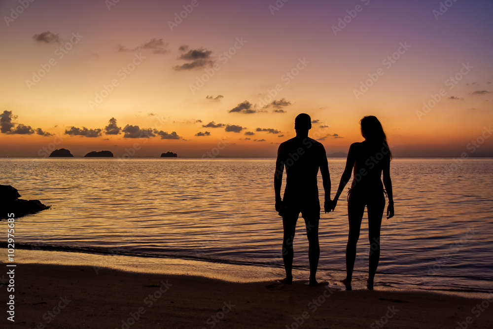 Young couple standing on the beach holding hands and watching tropical sunset