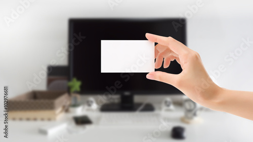 Hand holding and showing small blank white card on blurred working room background, horizontal style