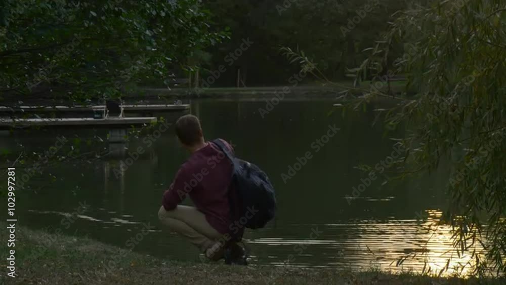 Man is Sitting at The River Lake Bank Man Tooks His Backpack On Man Has Stood Up and Walking Away Two Wooden Piers at River Lake Distantly Deck Chair Man is Sitting at The River Lake Bank Man Tooks His Backpack On Man Has Stood Up and Walking Away Two Wooden Piers at River Lake Distantly Deck Chair
