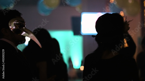 Two people within audience are dancing back to camera, man and woman moving to the music with masquerade masks on their faces at the special styled event. photo