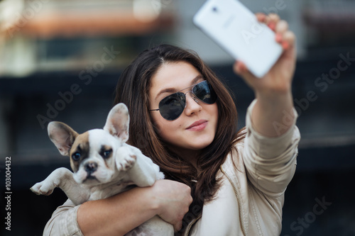 Beautiful young girl smiling and taking a selfie with her cute French bulldog puppy. Urban scene. Selective focus on girl. photo
