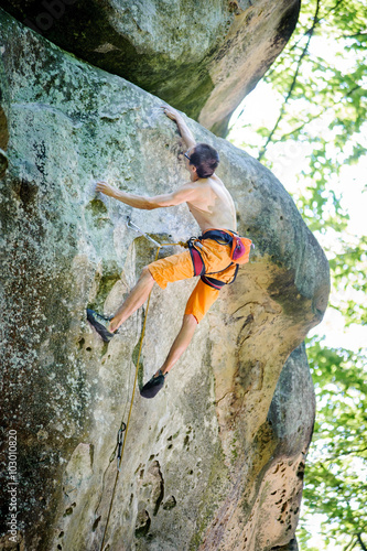 Athletic male climber climbing on a large boulder, with carbines and rope engaged. Outdoor summer day.