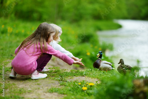 Two adorable sisters feeding ducks by a river Fototapet
