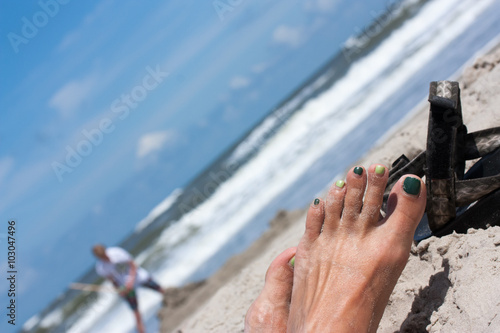 Nahaufnahme eines Fußes einer Frau mit grün lackierten Nägeln, im Hintergrund ein Kind, das eine Sandburg baut.