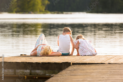 Traumhafter Sommer! Junge Frau sonnt sich auf einem Bootssteg und ihre beiden Kinder sitzen in der Sonne und beobachten etwas in Wasser. Schöne, entspannte Sommerstimmung, 