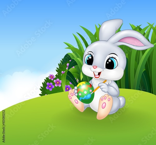 Cute Easter Bunny painting an egg on the Meadow background 