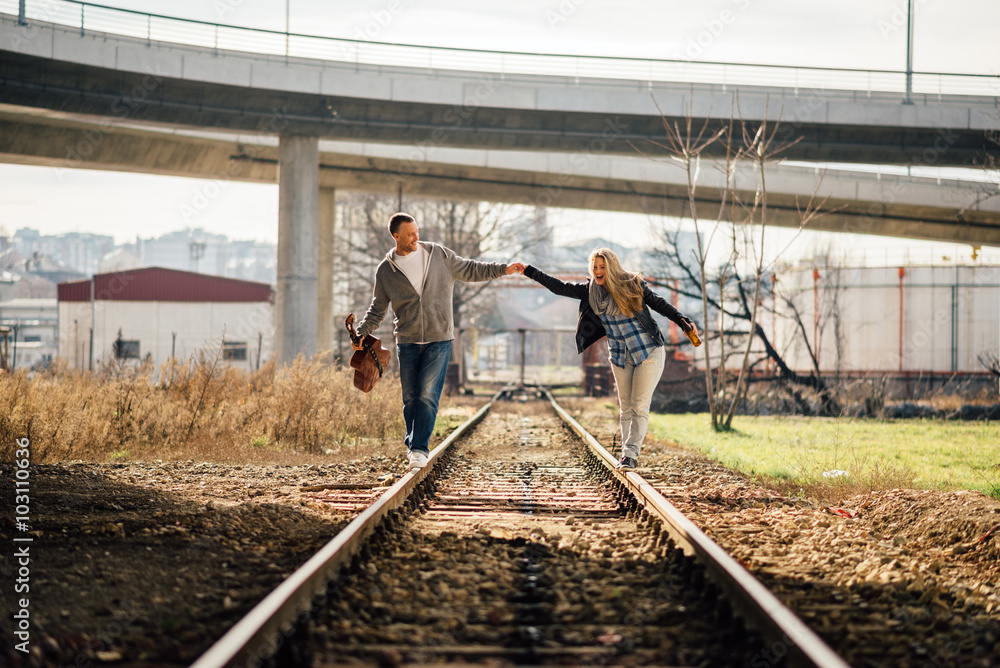 One couple, boy and girl, walking with guitar and beer on railway