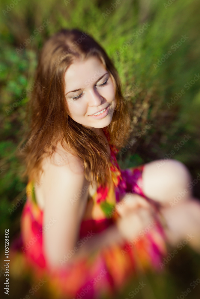 Naklejka premium Beautiful smiling ginger girl with closed eyes sitting on grass in summer park on sunny day. Selective focus on one eye, lensbaby blur effect. Relax and rest concept