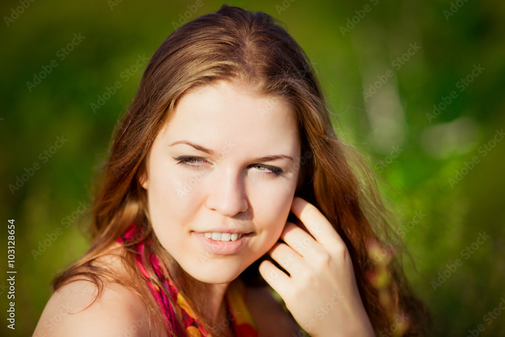 Close-up portrait of young woman with long red hair touching her face with her hand. Ginger girl sitting on grass in summer sunny field. Femininity concept