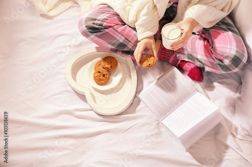 Woman in pajamas reading a book and drinking milk with cookies on her bed