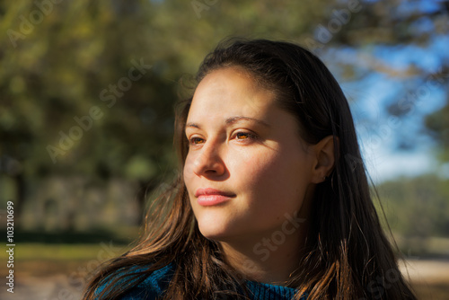 Portrait of a young white woman with brown hair and bright brown eyes outside in the sunlight looking contemplative thoughtful knowing unreadable unattainable
