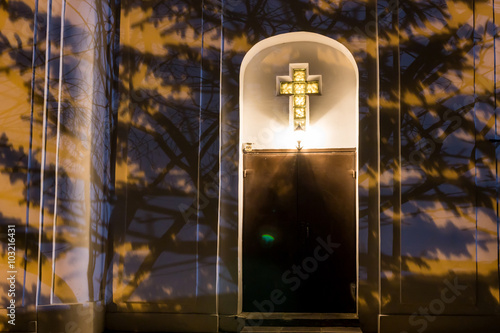 Christian Catholic cross on the facade of the church at night. Street lighting and tree branches create light shade. Widely
 photo