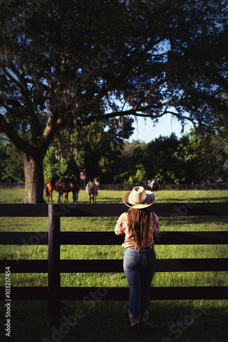 Cowgirl lady woman female wearing cowboy hat and flannel shirt with jeans leaning on country rural fence by a horse pasture paddock looking confident happy serene smart alone waiting watching patient