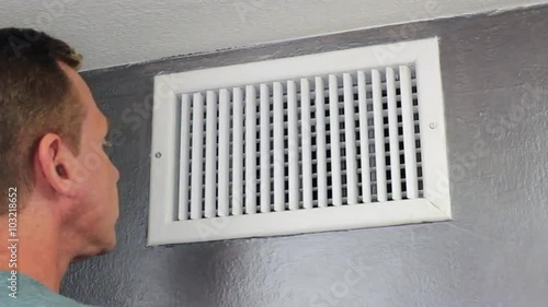 Man Examining an Air Vent. Mature male peering inside an upper wall white grid air duct on a silver wall near a white ceiling. A guy inspecting a heating and cooling air register duct for maintenance.