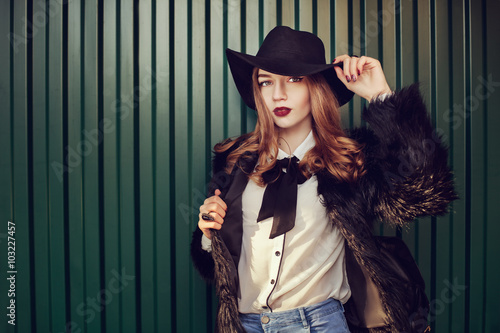 Street closeup portrait of a young beautiful lady wearing stylish black fur coat and wide-brimmed hat. Model looking at camera. Female fashion concept. Dark green background. Toned