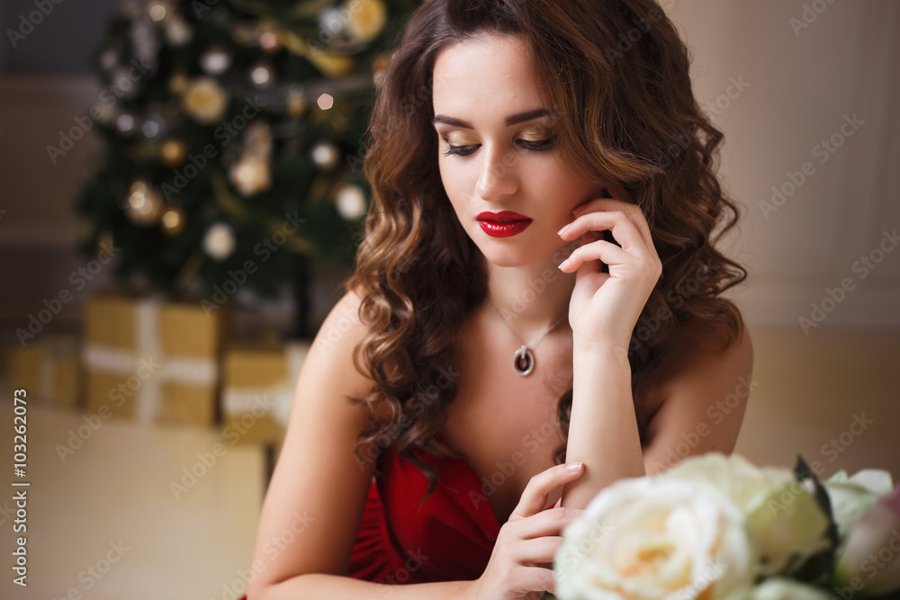 Close-up portrait of beautiful young woman in gorgeous red velvet evening dress sitting by the table in expensive interior