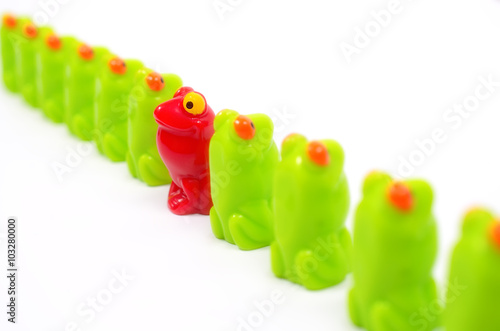 Line of green toy frogs. Among them, one red frog. Close up view. Different from the others. At home among strangers. Concept of antagonism.